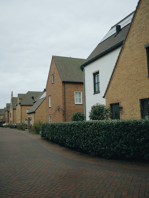 A quiet residential street featuring modern terraced homes with varying brick facades, some painted white, and pitched roofs with slate tiles. The front gardens are neatly maintained, separated by wel